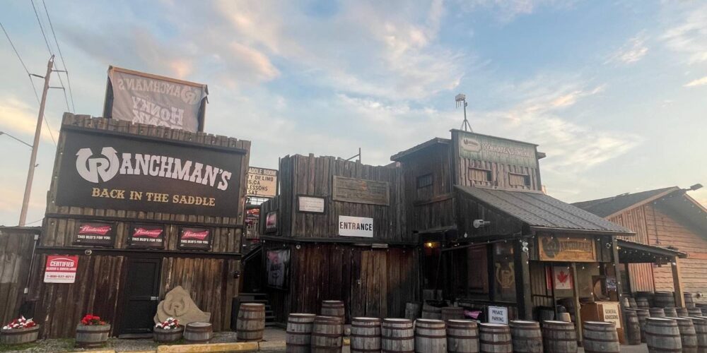 The rustic exterior of Ranchman’s Cookhouse and Dance Hall, with wooden barrels, a Canadian flag, and signage indicating “Entrance” and “Private Parking.”