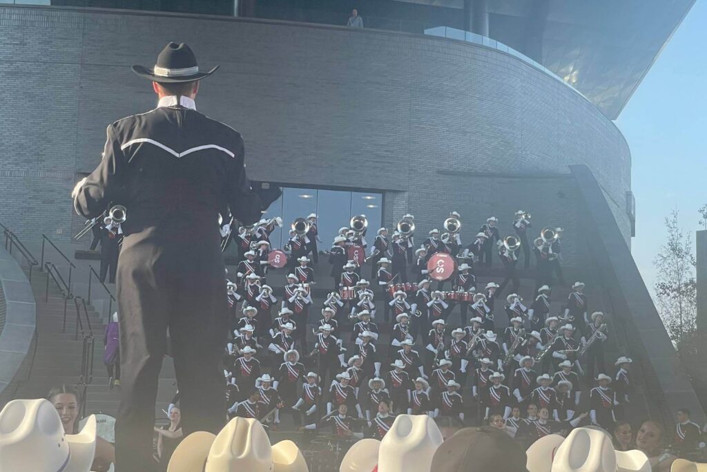 The Calgary Stampede band performing on the steps outside of the BMO Centre with the conductor dressed in black, facing away from the camera, leading the ensemble. The band is wearing cowboy hats and "CS" initials are on the bass drums. The musicians are dressed in coordinated uniforms with white cowboy hats, and the scene captures the grandeur of a formal band performance.