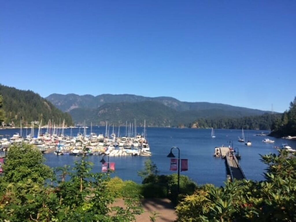 A scenic view of Deep Cove in North Vancouver, BC, Canada, showcasing a bustling marina filled with boats, surrounded by lush mountains and a clear blue sky.