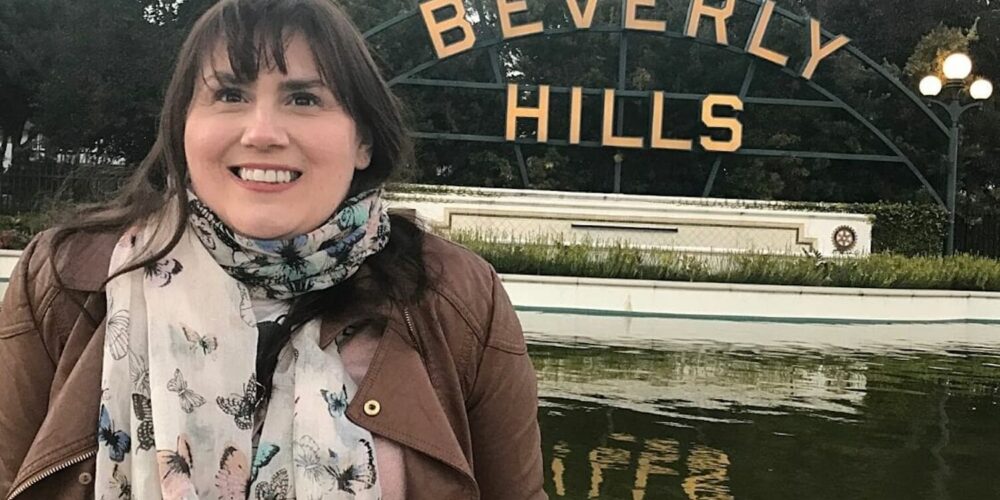 A cheerful woman, Gemma Lawrence, with dark hair, wearing a brown jacket and a butterfly-print scarf, stands before the iconic Beverly Hills sign with a pond and greenery in the foreground.