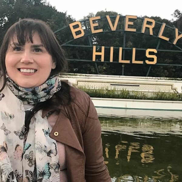 A cheerful woman, Gemma Lawrence, with dark hair, wearing a brown jacket and a butterfly-print scarf, stands before the iconic Beverly Hills sign with a pond and greenery in the foreground.