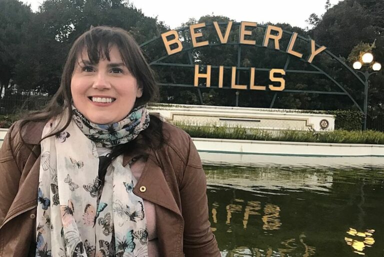 A cheerful woman, Gemma Lawrence, with dark hair, wearing a brown jacket and a butterfly-print scarf, stands before the iconic Beverly Hills sign with a pond and greenery in the foreground.