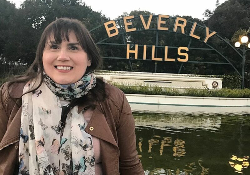 A cheerful woman, Gemma Lawrence, with dark hair, wearing a brown jacket and a butterfly-print scarf, stands before the iconic Beverly Hills sign with a pond and greenery in the foreground.