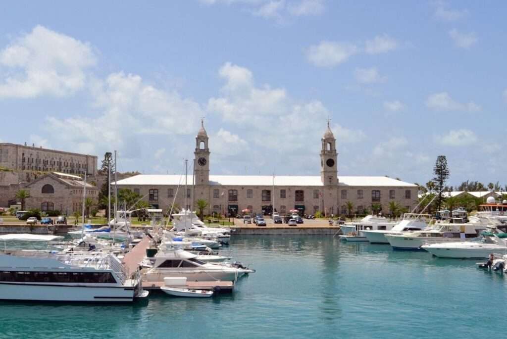 The historic Royal Naval Dockyard in Bermuda, showcasing its iconic clock tower and rugged limestone buildings, overlooks a serene marina dotted with various boats under a clear blue sky.