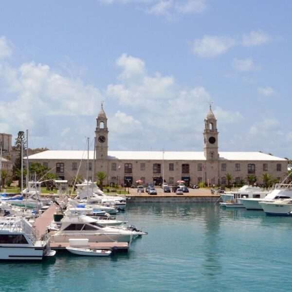 The historic Royal Naval Dockyard in Bermuda, showcasing its iconic clock tower and rugged limestone buildings, overlooks a serene marina dotted with various boats under a clear blue sky.