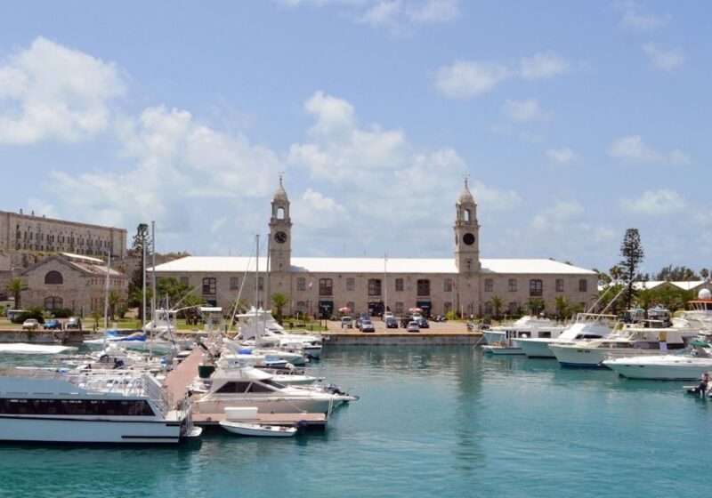 The historic Royal Naval Dockyard in Bermuda, showcasing its iconic clock tower and rugged limestone buildings, overlooks a serene marina dotted with various boats under a clear blue sky.