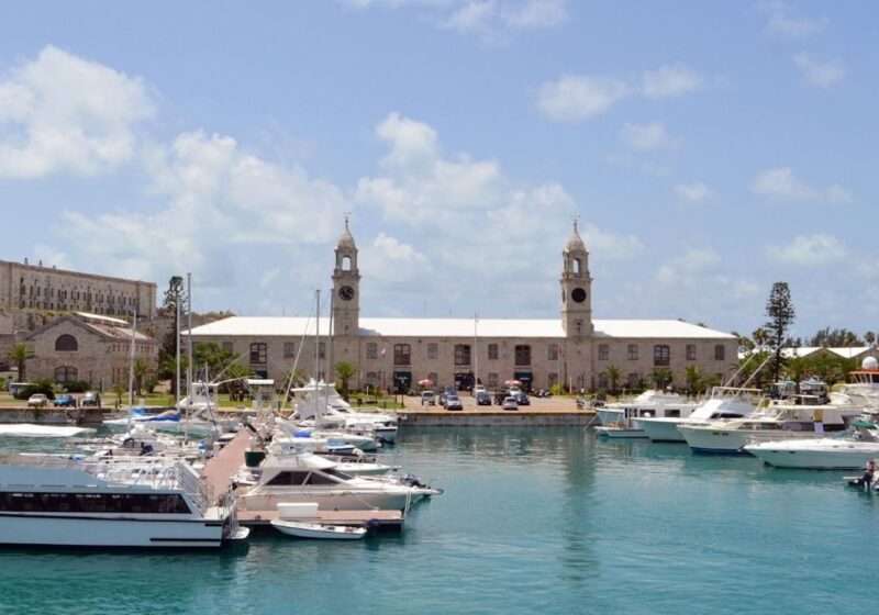 The historic Royal Naval Dockyard in Bermuda, showcasing its iconic clock tower and rugged limestone buildings, overlooks a serene marina dotted with various boats under a clear blue sky.