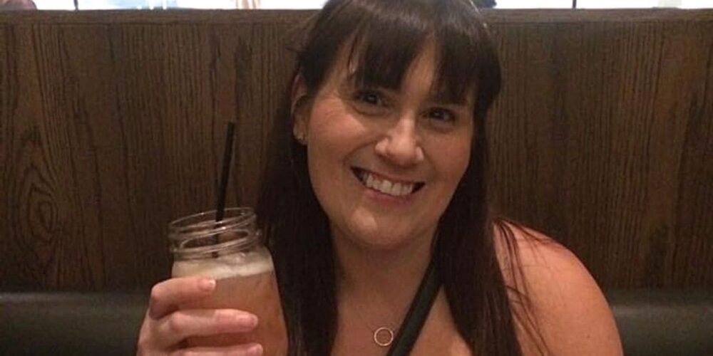 A smiling woman, Gemma Lawrence, with dark hair, wearing a white top and a necklace with a circular pendant, holding a mason jar glass with a dark straw, seated in a restaurant with a wooden booth backdrop.