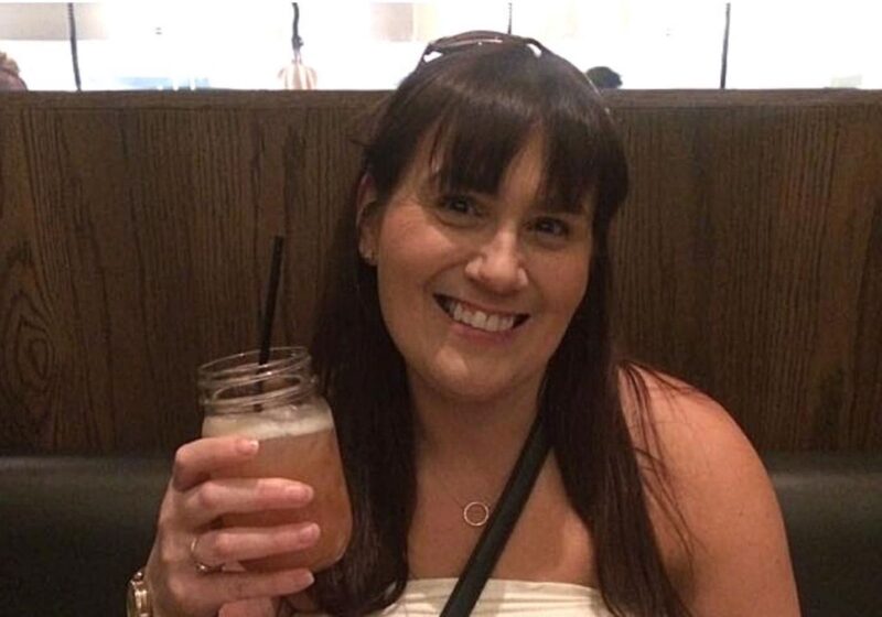 A smiling woman, Gemma Lawrence, with dark hair, wearing a white top and a necklace with a circular pendant, holding a mason jar glass with a dark straw, seated in a restaurant with a wooden booth backdrop.