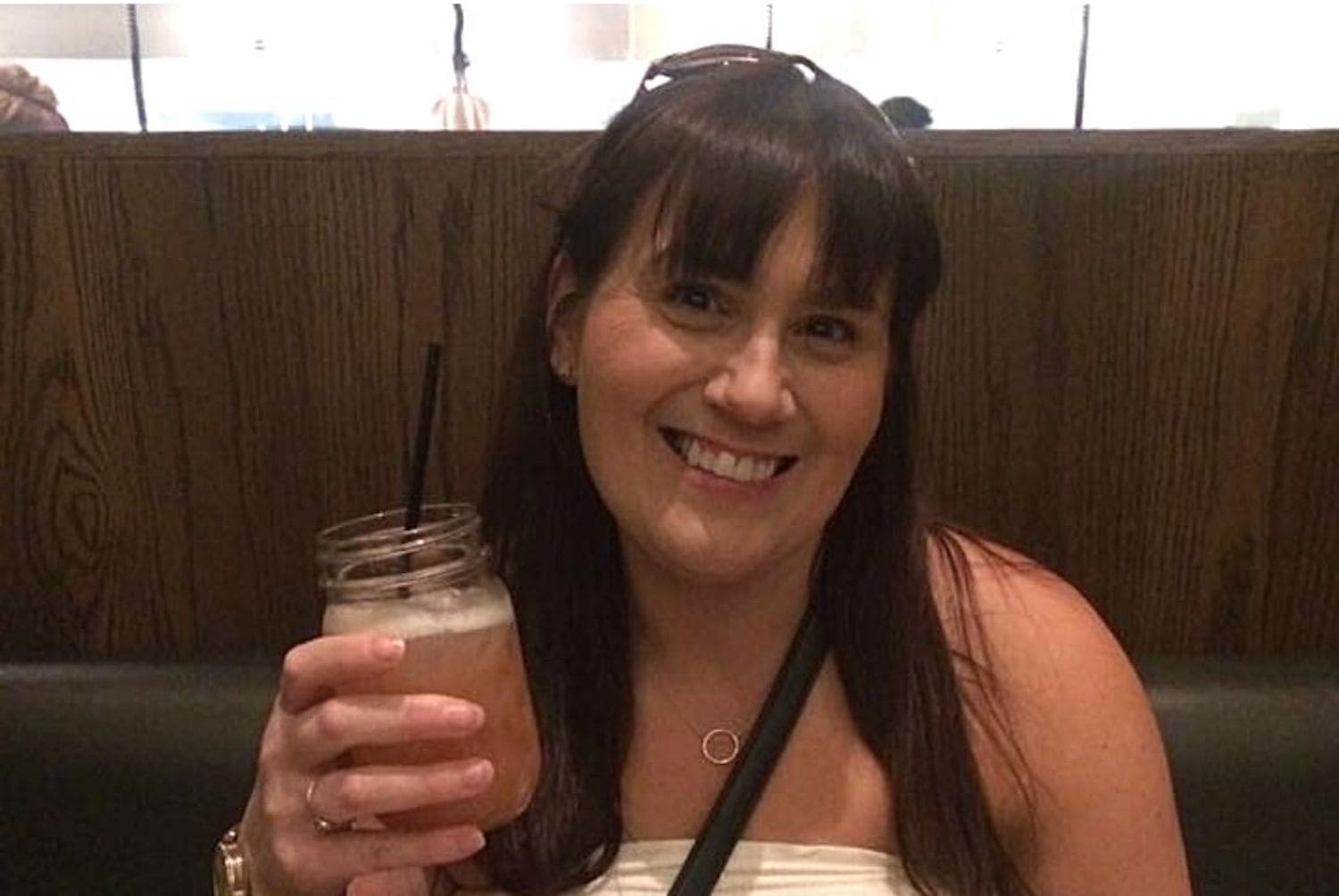 A smiling woman, Gemma Lawrence, with dark hair, wearing a white top and a necklace with a circular pendant, holding a mason jar glass with a dark straw, seated in a restaurant with a wooden booth backdrop.