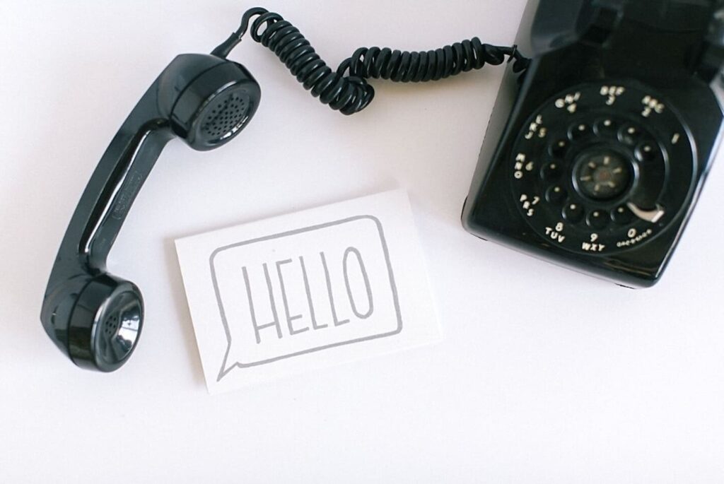 Vintage black rotary phone with the receiver off the hook, next to a card with 'HELLO' printed on it, set against a white background.