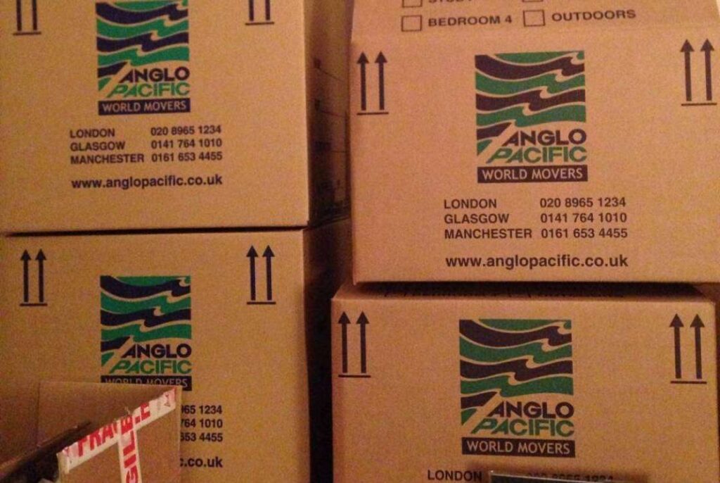 Stack of cardboard moving boxes with 'Anglo Pacific World Movers' branding, contact information for London, Glasgow, and Manchester, and the company's website, indicating a moving or relocation service.
