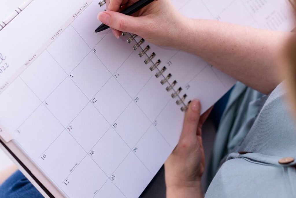 Close-up of a person's hands writing in a planner with a black pen, focusing on a clean calendar page, suggesting organization and schedule planning.