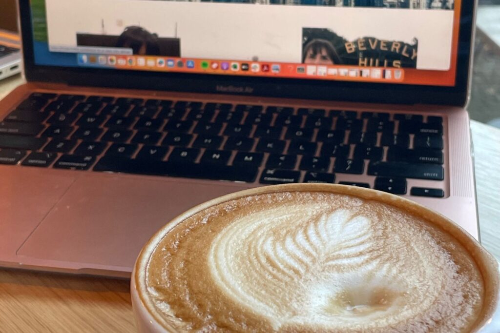 A close-up of a frothy coffee cup with a detailed latte art design, sitting in front of an open laptop. The screen displays a webpage, with part of a "Beverly Hills" sign visible, suggesting a casual workspace. setting.