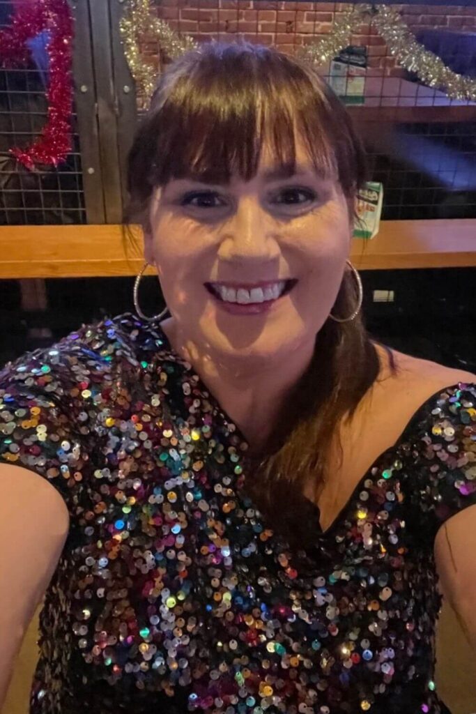 A woman, Gemma Lawrence, taking a selfie while wearing a colorful, sequined top in a dimly lit bar or club. The background features some festive decorations and a wooden interior.