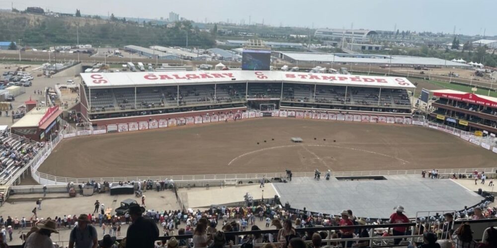 A wide shot of the Calgary Stampede rodeo arena from a high vantage point, with the audience filling the stands and the rodeo grounds below.