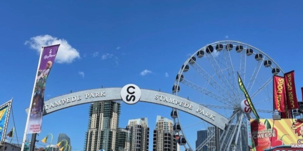 A bright daytime view of a large archway that reads "Stampede Park." A Ferris wheel and various food stalls are visible, with a clear blue sky above.