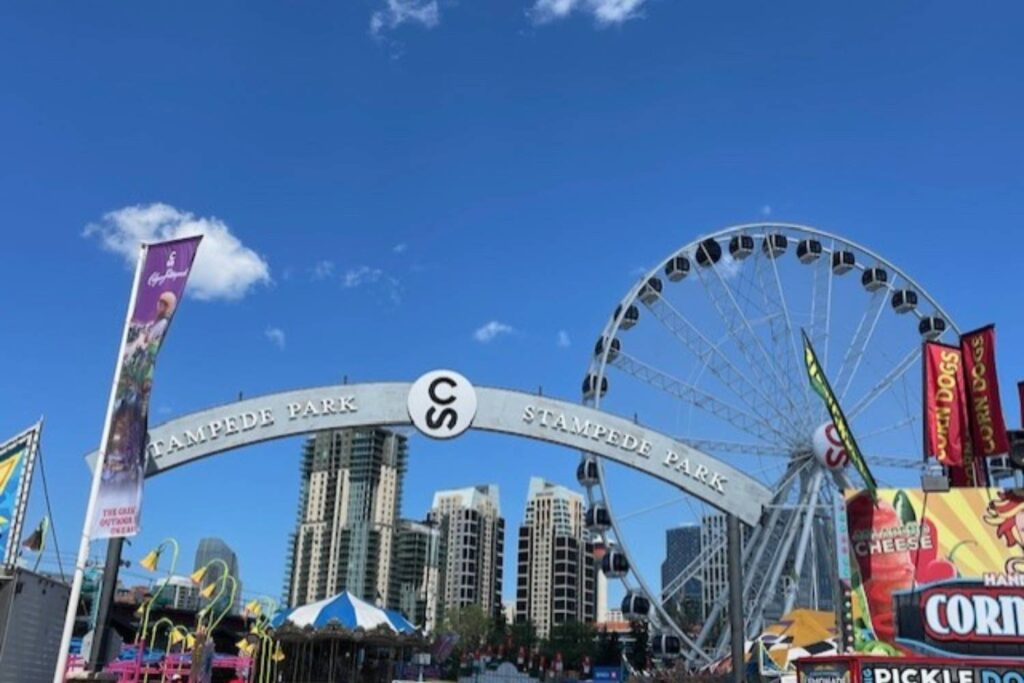 A bright daytime view of a large archway that reads "Stampede Park." A Ferris wheel and various food stalls are visible, with a clear blue sky above.