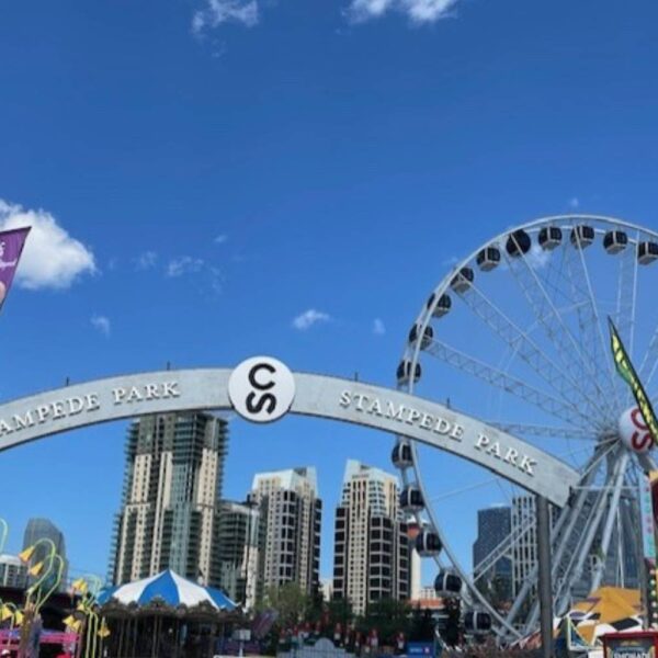 A bright daytime view of a large archway that reads "Stampede Park." A Ferris wheel and various food stalls are visible, with a clear blue sky above.