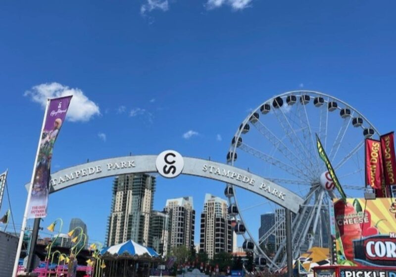 A bright daytime view of a large archway that reads "Stampede Park." A Ferris wheel and various food stalls are visible, with a clear blue sky above.