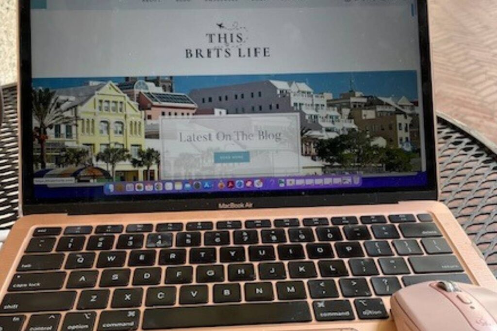 A close-up of a pink MacBook Air, showing the "This Brit's Life" website featuring a colorful street scene with pastel buildings. The screen highlights the "Latest On The Blog" section. A pink wireless mouse is positioned next to the laptop on a metal outdoor table.