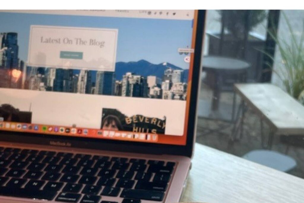 A partial view of a MacBook Air displaying a website with a scenic city skyline, likely Vancouver, with the text "Latest On The Blog" in a prominent box. The scene is set in a cozy café with an empty table and plant in the background.