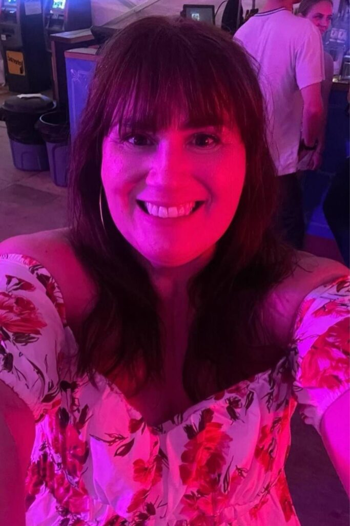 Gemma Lawrence of This Brits Life smiling brightly in a pink-lit environment wearing a floral dress, with other people and a bar in the background enjoying herself in the Ranchman's Stampede Tent.