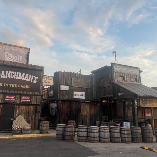 The rustic exterior of Ranchman’s Cookhouse and Dance Hall, with wooden barrels, a Canadian flag, and signage indicating “Entrance” and “Private Parking.”