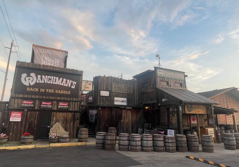 The rustic exterior of Ranchman’s Cookhouse and Dance Hall, with wooden barrels, a Canadian flag, and signage indicating “Entrance” and “Private Parking.”