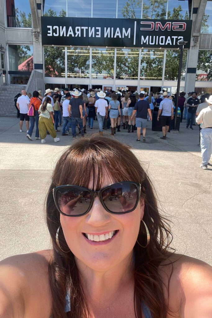 Gemma Lawrence wearing sunglasses and a blue tank top takes a selfie in front of the "GMC Stadium Main Entrance." on Stampede Park in Calgary. Behind her, a large crowd, many in cowboy hats, is entering the venue.