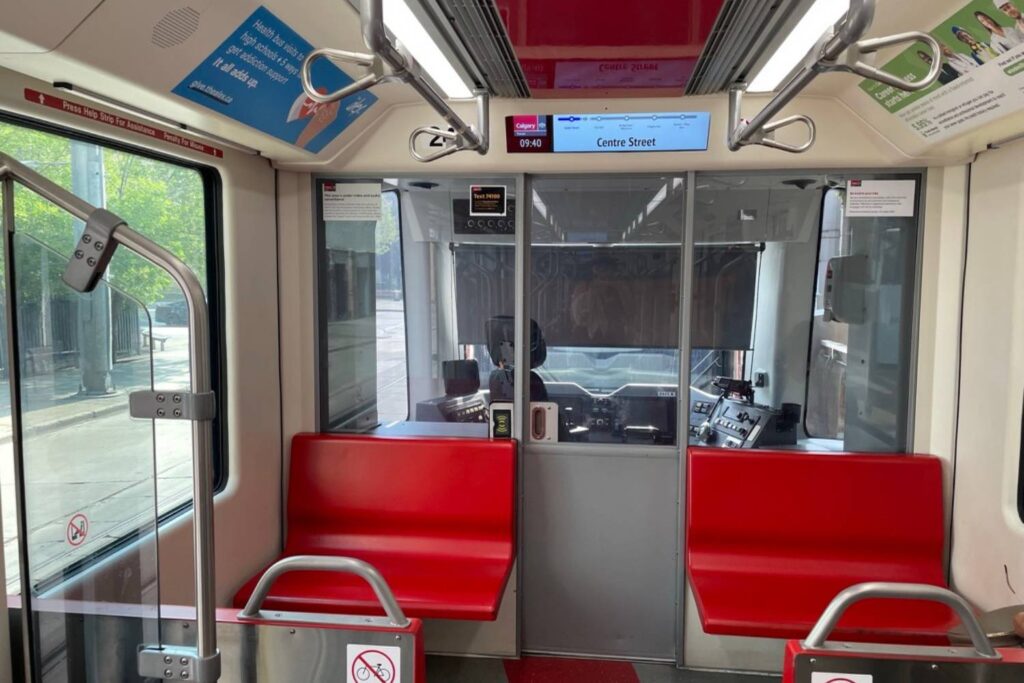 The interior of a Calgary C-Train car, focusing on the area near the front of the train. The setup includes red plastic seats on either side of the aisle and a glass partition separating the passenger area from the driver's cab. A digital display above the partition shows the current station as "Centre Street" and the time as 09:40. The environment is clean and well-lit, with signs posted on the walls providing information and instructions to passengers.