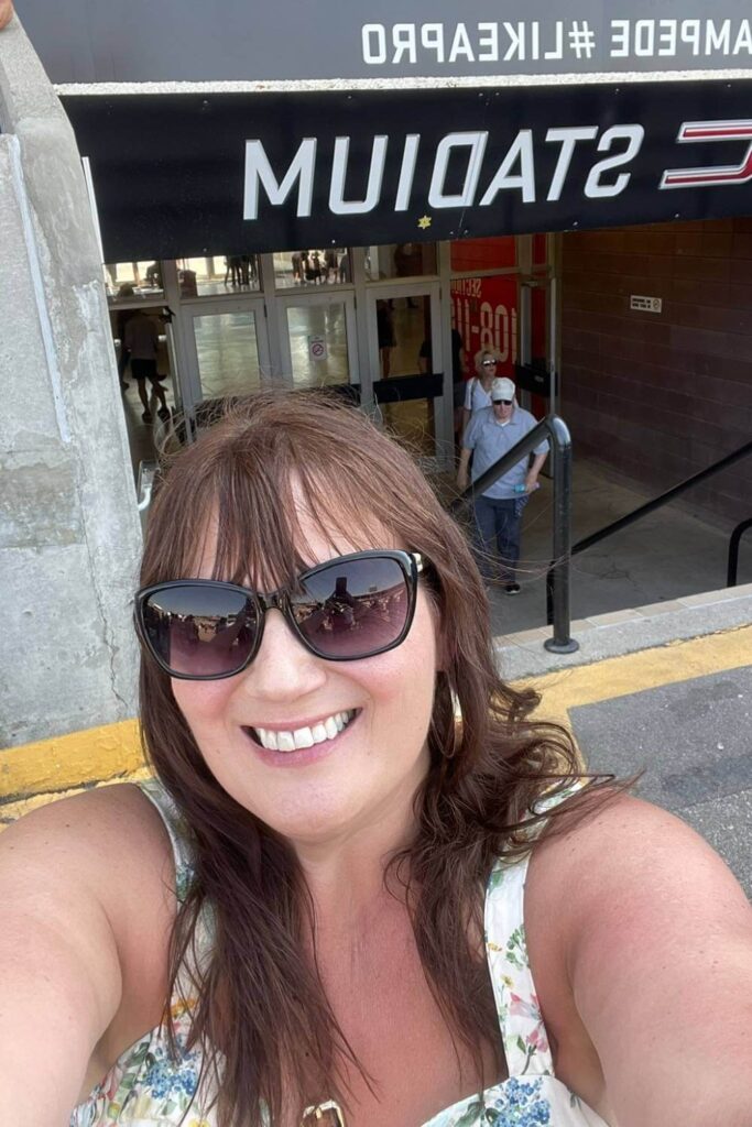 Gemma Lawrence smiling while wearing sunglasses taking a selfie inside the GMC Stadium during the rodeo with the stadium sign partially visible above her.