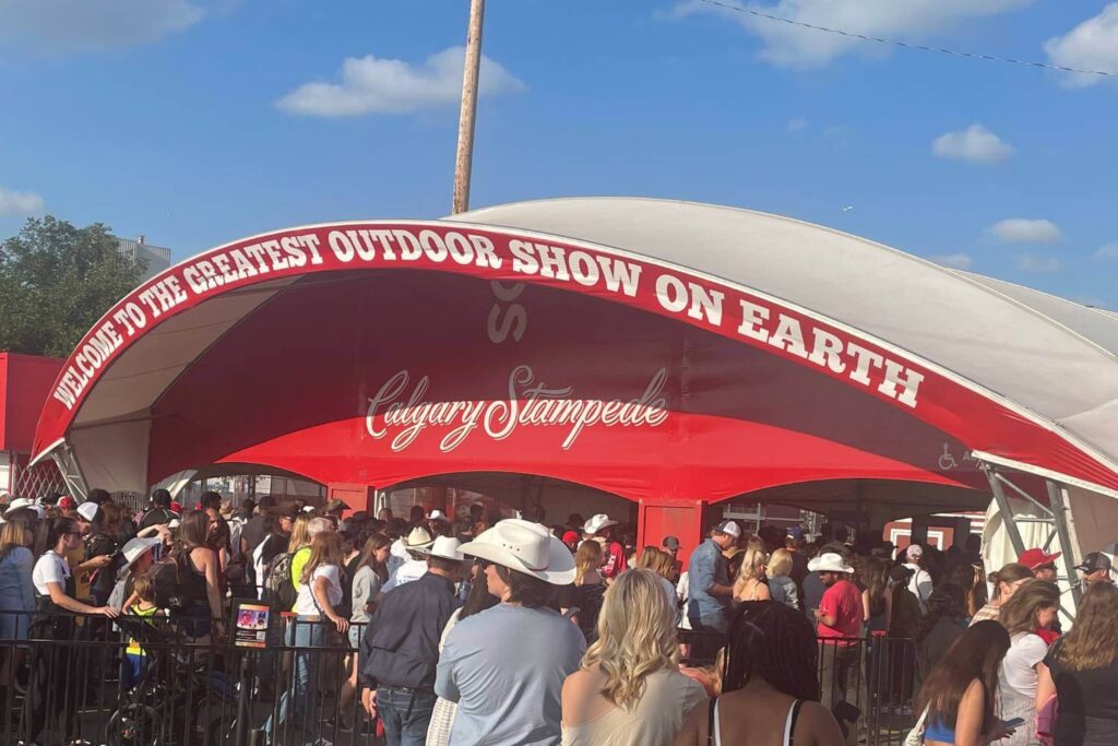 An entrance arch at the Calgary Stampede that reads "Welcome to the Greatest Outdoor Show on Earth," with a crowd of attendees waiting to enter the event.