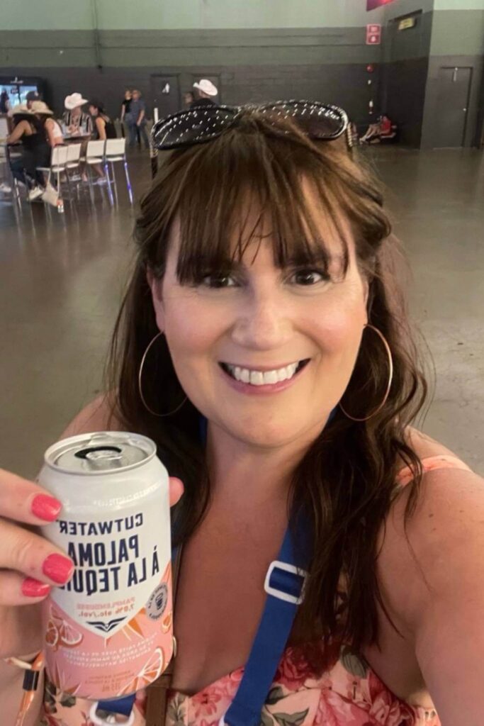 Gemma Lawrence in a pink floral dress with sunglasses on her head holding a can of Cutwater Paloma Tequila, smiling inside the Big Four Roadhouse during the Calgary Stampede with other attendees in the background.