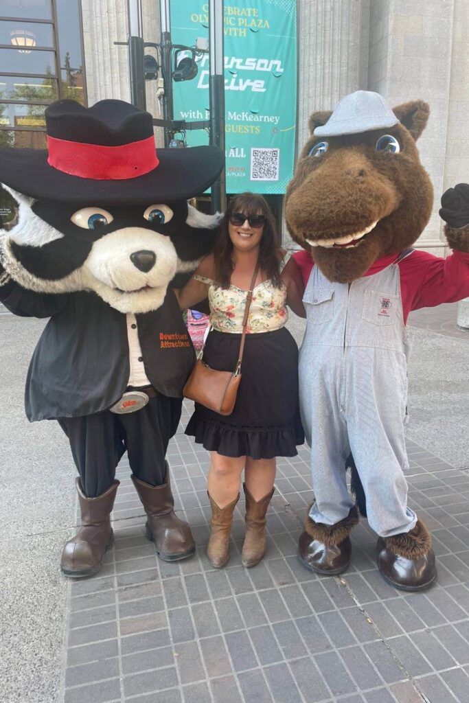 Gemma Lawrence posing with two mascots at the Flour Rope Square pancake breakfast during the Calgary Stampede—one dressed as a raccoon and the other as a beaver—against an urban backdrop of downtown Calgary, Alberta.