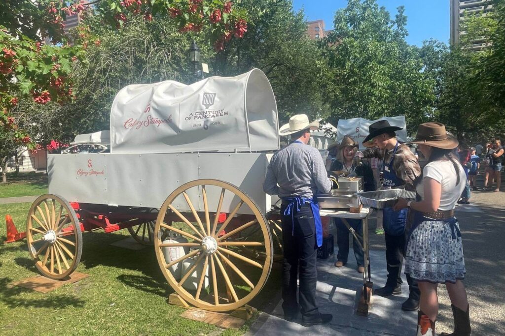 A group of people dressed in western attire, including cowboy hats, serve pancakes from a traditional chuckwagon setup during an outdoor event surrounded by trees.