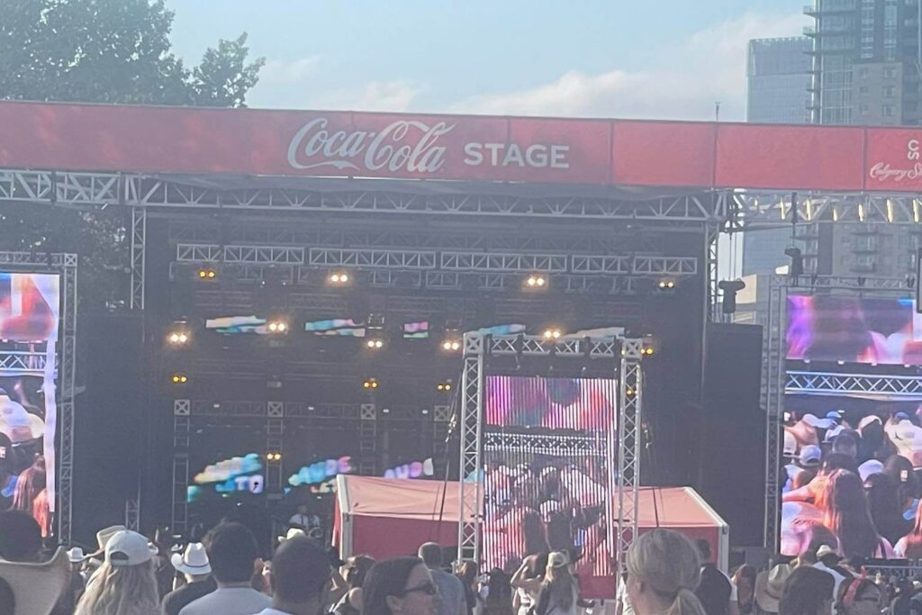 A view of the Coca-Cola Stage at the Calgary Stampede, with concert lighting and large video screens set up for a live performance. A crowd of people, many in cowboy hats, gather to watch.