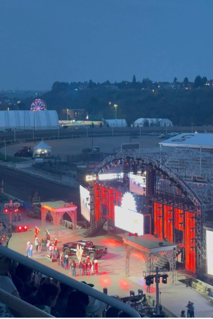 A large outdoor stage lit up with red and white lights during the evening event at the Calgary Stampede. Performers stand in front of a pickup truck, while a Ferris wheel and white event tents are visible in the distance under a darkening sky.