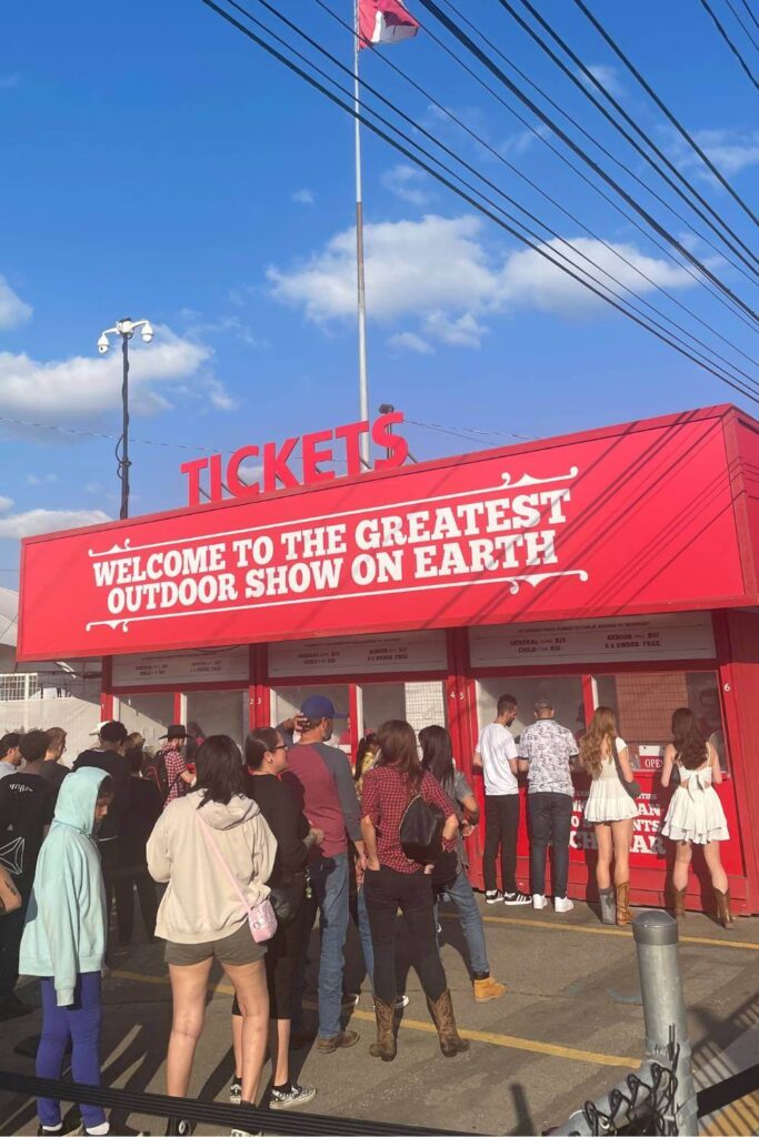 A red ticket booth at the Calgary Stampede labeled "Welcome to the Greatest Outdoor Show on Earth," with a line of attendees waiting to purchase tickets.