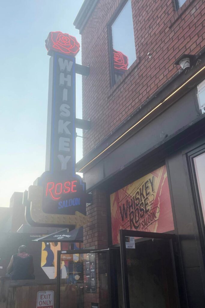 The neon sign for "Whiskey Rose Saloon" against the brick exterior of the building, with a prominent red rose design and blue and red lettering.