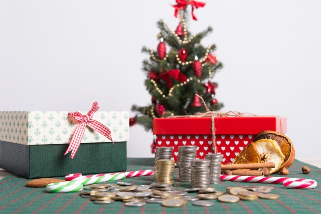 Stacks of coins and festive gift boxes on a table in front of a decorated Christmas tree. The scene includes candy canes, dried orange slices, and cinnamon sticks, symbolizing holiday budgeting and spending.