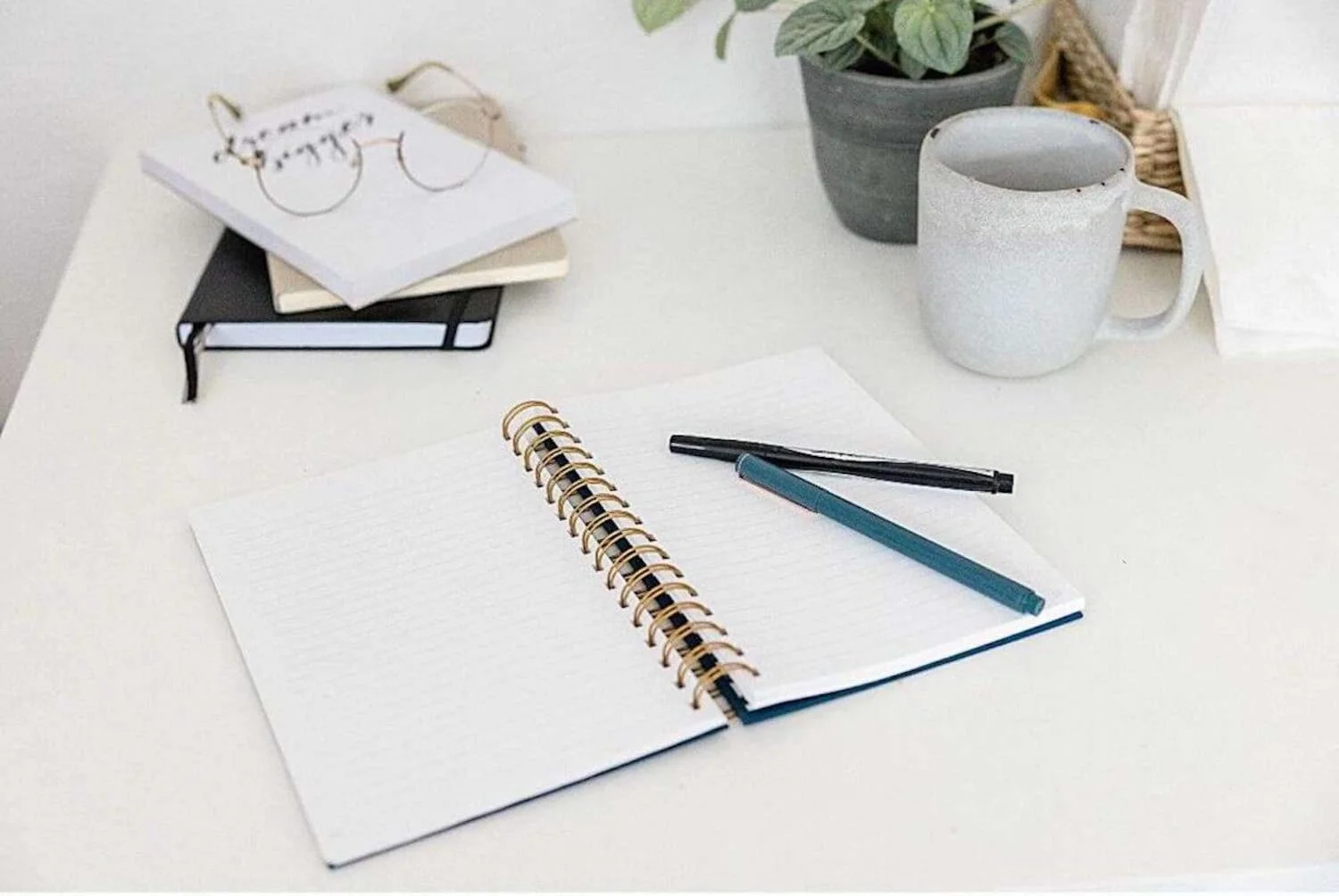 A minimalist desk setup with an open spiral notebook, two pens, and a stack of books beside a potted plant and a textured ceramic mug.