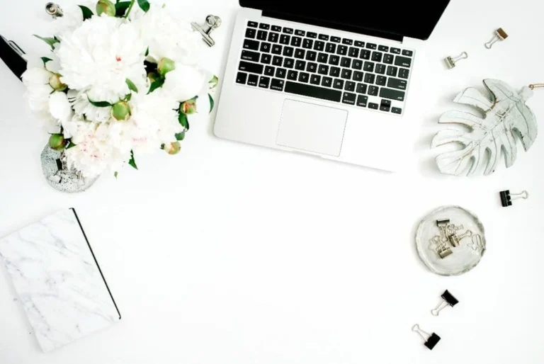 A minimalist workspace setup with a bouquet of white flowers in a vase, a laptop, a marble-patterned notebook, a metal palm leaf decoration, and assorted stationery items neatly arranged on a white surface, creating an aesthetic and organized look ideal for a blogging environment.