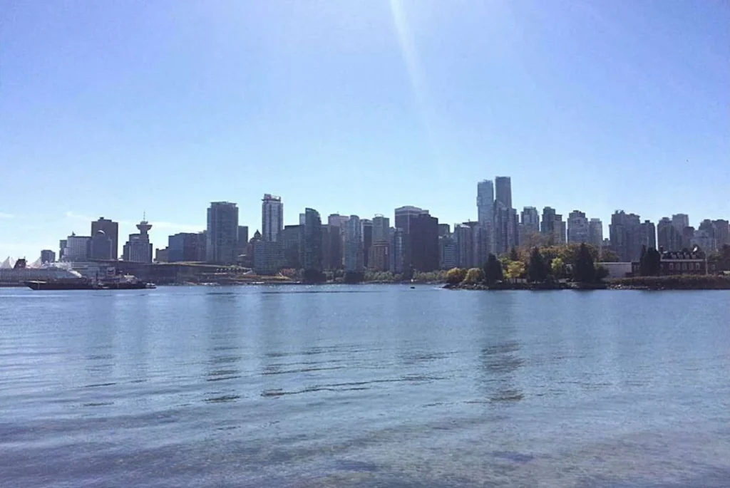A view across the calm waters towards the dense urban skyline of Vancouver, with clear skies above and a hint of greenery at the water's edge.