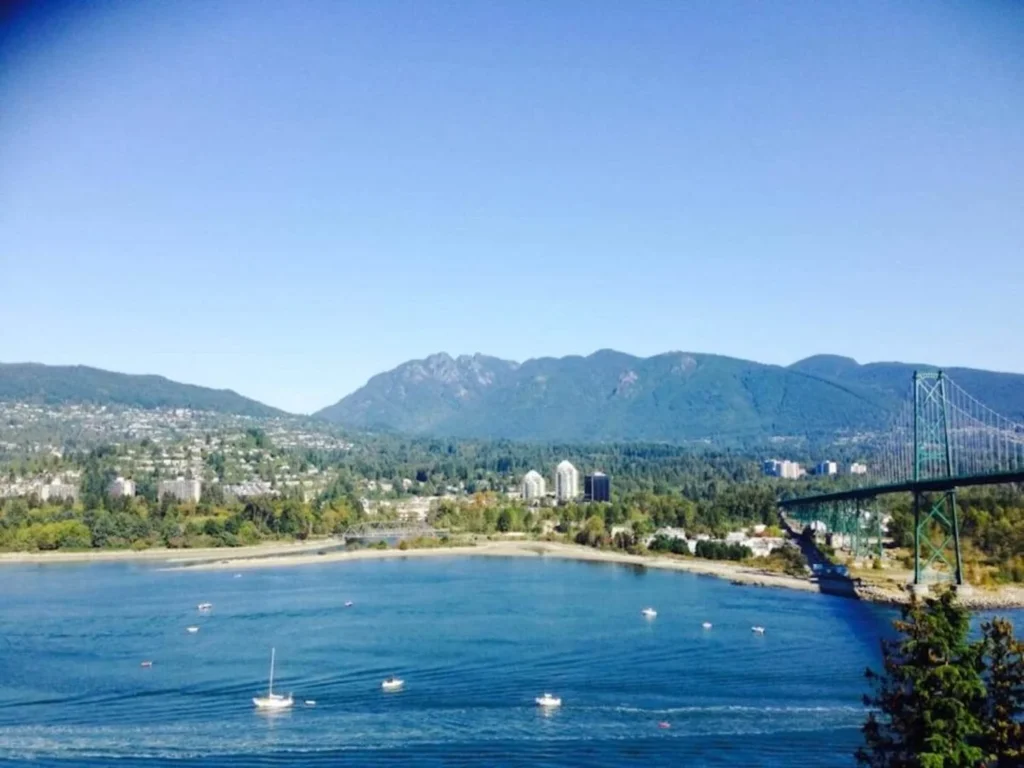 A panoramic view from Prospect Point in Stanley Park, Vancouver, showcasing the Lions Gate Bridge, a calm bay with boats, and a backdrop of mountains under a clear blue sky.