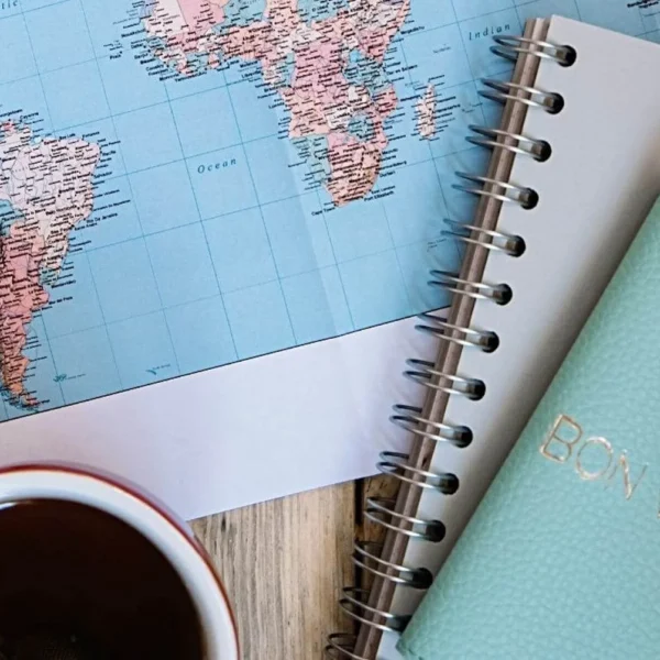 Top view of a travel planning setup with a world map focused on South America, a spiral-bound notebook, a cup of tea, and a mint green passport holder with 'BON VOYAGE' embossed on it.
