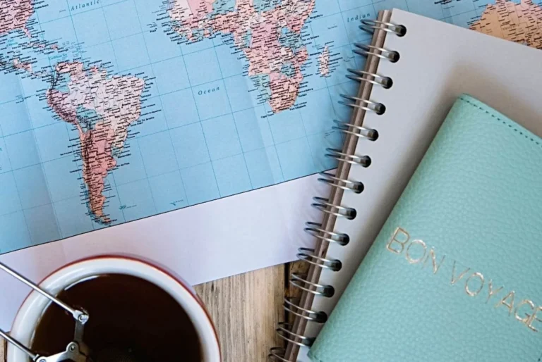 Top view of a travel planning setup with a world map focused on South America, a spiral-bound notebook, a cup of tea, and a mint green passport holder with 'BON VOYAGE' embossed on it.