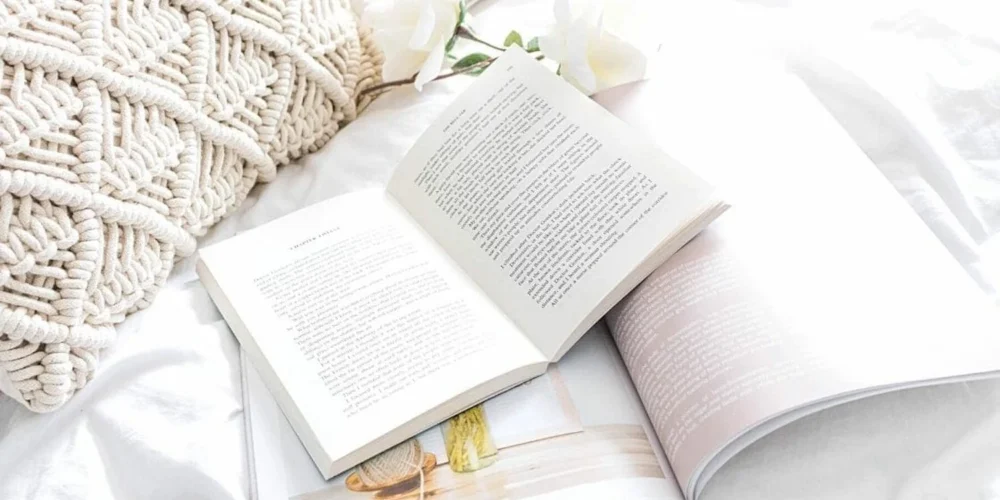 A cozy reading nook captured in a high-key photograph with a soft knitted throw pillow, an open book with visible text, and delicate white flowers resting on a crumpled white bedsheet, evoking a serene and comfortable atmosphere perfect for leisurely reading.