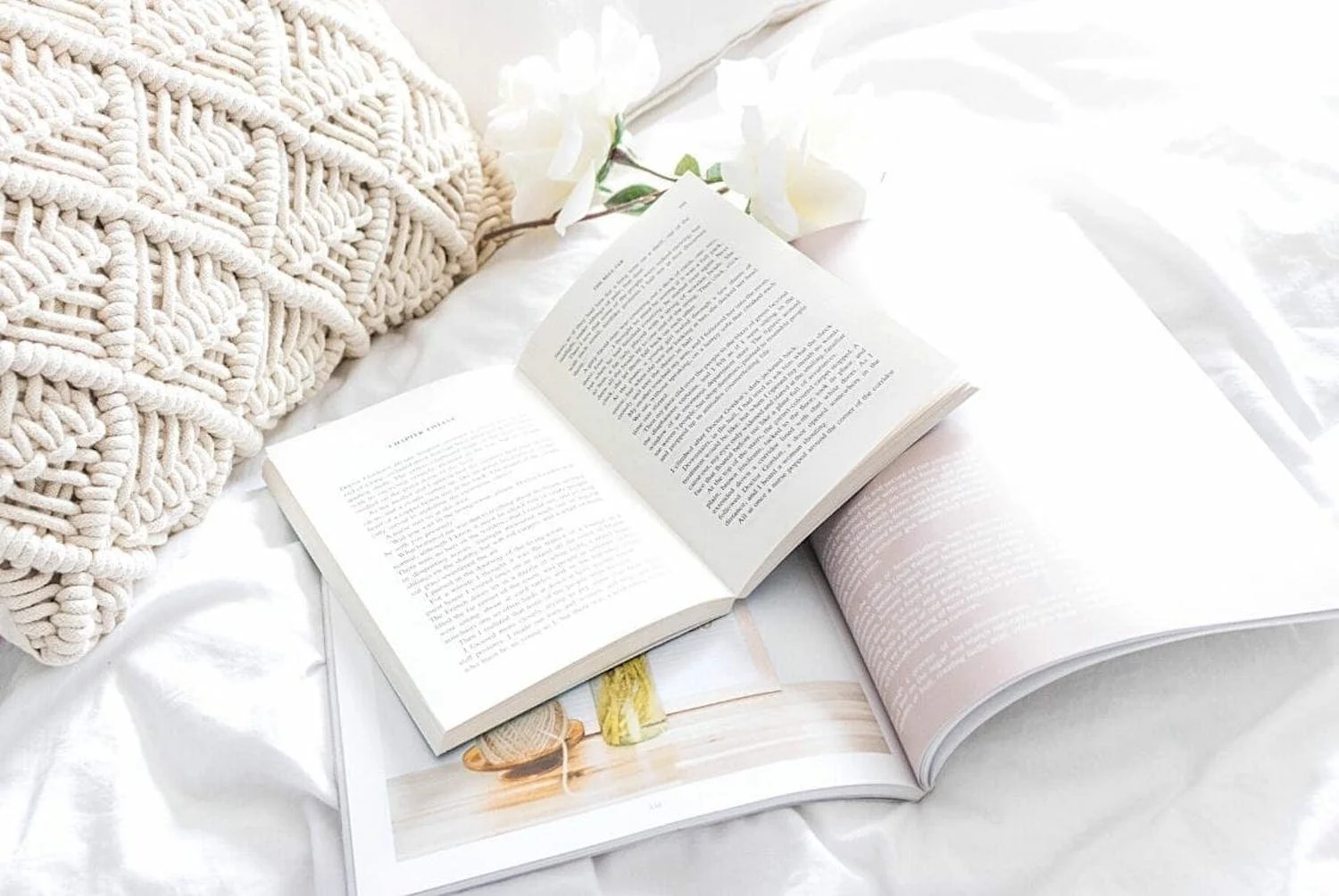 A cozy reading nook captured in a high-key photograph with a soft knitted throw pillow, an open book with visible text, and delicate white flowers resting on a crumpled white bedsheet, evoking a serene and comfortable atmosphere perfect for leisurely reading.
