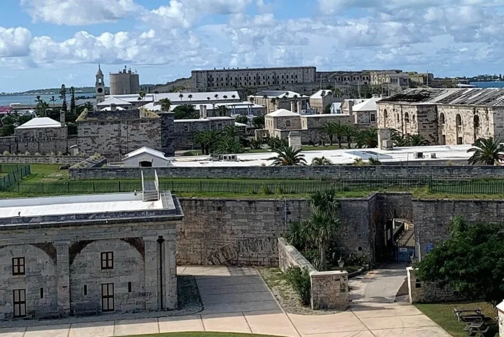 An aerial view of the historic Royal Naval Dockyard in Bermuda, showcasing the stone fortifications and maritime buildings with lush green lawns, under a partly cloudy sky. The architecture reflects Bermuda's colonial and maritime heritage.